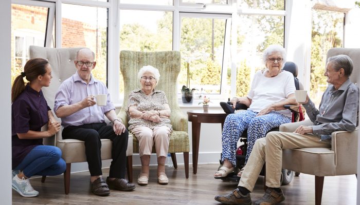 Male And Female Residents Sitting In Chairs And Talking With Carer In Lounge Of Retirement Home